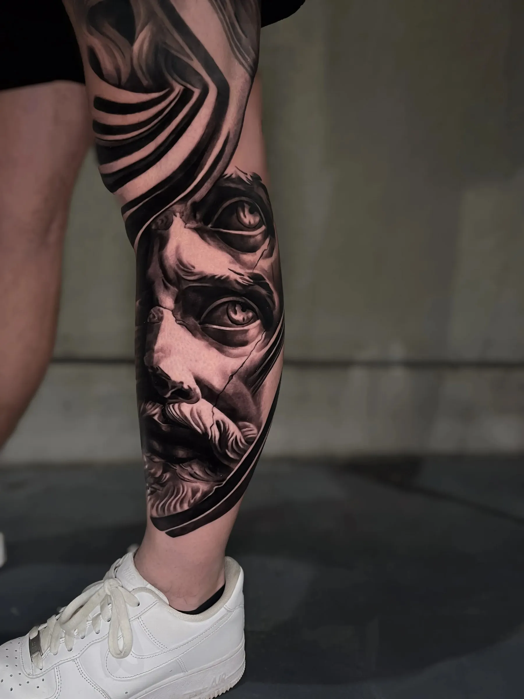 Illusion with real form: klassiek Grieks beeldengezicht in zwart-grijs met abstracte zwarte en grijze vormen eromheen, op het onderbeen